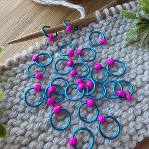 Pu&ograve; includere: Segnapunti turchesi con perline rosa su una superficie a maglia beige. Un ferro da maglia in legno e un gomitolo di lana suggeriscono un tema artigianale. Per progetti di maglieria.