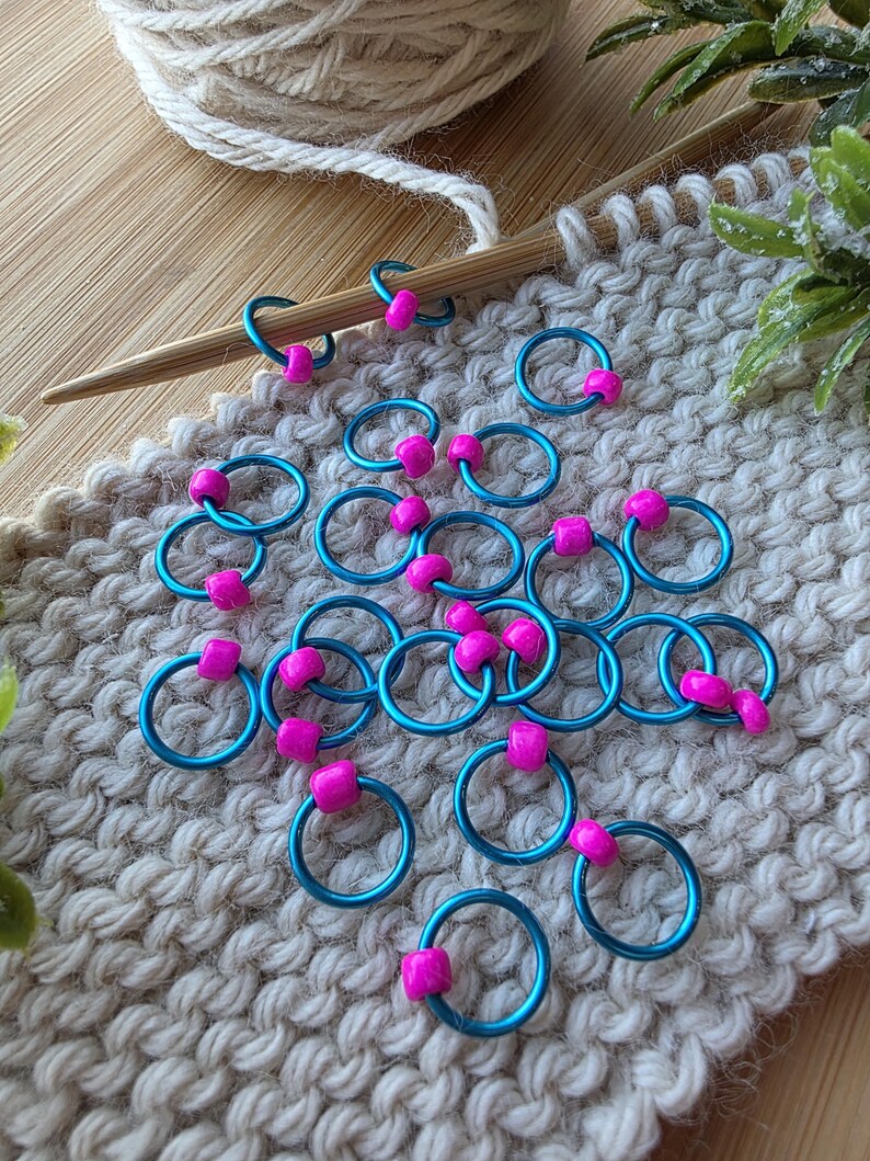 Pu&ograve; includere: Segnapunti turchesi con perline rosa su una superficie a maglia beige. Un ferro da maglia in legno e un gomitolo di lana suggeriscono un tema artigianale. Per progetti di maglieria.