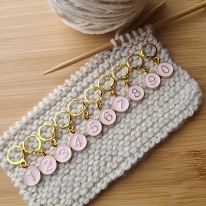 May include: Nine gold stitch markers with pink numbered circles. The numbers 1 through 9 are visible on the markers. The markers are on a beige knitted fabric.