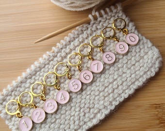 May include: Nine gold stitch markers with pink numbered circles. The numbers 1 through 9 are visible on the markers. The markers are on a beige knitted fabric.