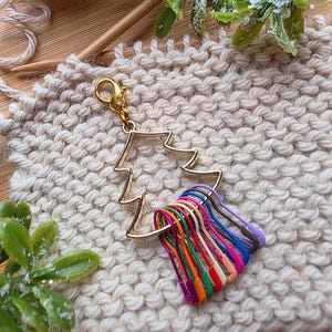 May include: A gold-toned metal Christmas tree-shaped charm featuring a lobster clasp at the top. Attached to the base of the tree are several small, colorful metal safety pin-style stitch markers in shades of purple, blue, pink, orange, and green.