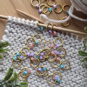May include: Gold-colored stitch markers with pastel beads in pink, blue, and purple. The markers are on a knitted swatch with wooden knitting needles and a ball of yarn. Ideal for knitting and crochet projects.