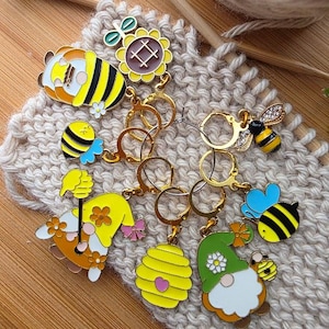 May include: A collection of colorful bee and gnome-themed stitch markers. The markers feature various designs, including bees, sunflowers, and gnomes, with gold-colored metal rings. The markers are set on a knitted background.