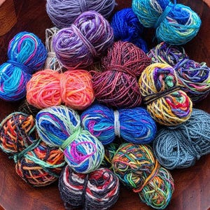 May include: A wooden bowl filled with colorful yarn skeins. The yarn is in various shades of blue, purple, orange, and green. The skeins are tightly wound and ready for crafting.