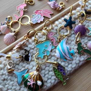 May include: A collection of colorful sea-themed stitch markers, including a mermaid, a starfish, a seashell, a seahorse, a shark, a fish, and a whale. The markers are made of metal and have a gold finish.