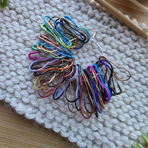 May include: A collection of colorful stitch markers in various hues, including blue, green, pink, and gold. The markers are arranged on a cream-colored knitted surface, with a wooden knitting needle visible in the background. These are essential tools for knitting and crochet projects.