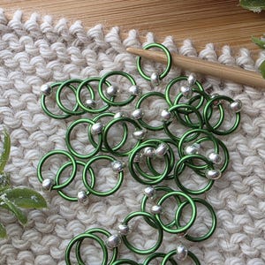 May include: A collection of green metal stitch markers with silver beads, arranged on a cream-colored knitted surface. A wooden knitting needle rests nearby. These markers are used for knitting and crochet projects.