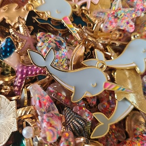 May include: A collection of colorful charms featuring a variety of designs, including narwhals, mermaids, seashells, and stars. The charms are made of metal and have a gold finish. They are perfect for adding a touch of whimsy to jewelry, bags, or other accessories.
