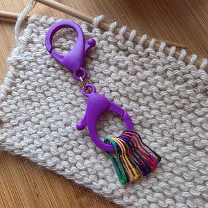 May include: A purple clasp with a ring of colorful stitch markers rests on a knitted swatch. The knitting project is made of a light beige yarn, with wooden knitting needles and a ball of yarn in the background. The stitch markers are in various colors.