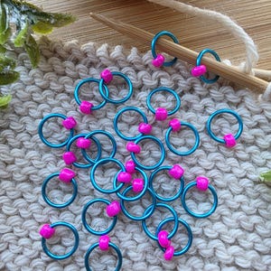 Pu&ograve; includere: Segnapunti color turchese con perline rosa sparsi su una superficie lavorata a maglia. Due segnapunti sono su un ferro da maglia di legno. Un gomitolo di lana e del verde sono sullo sfondo. Questi segnapunti sono per progetti di maglia e uncinetto.