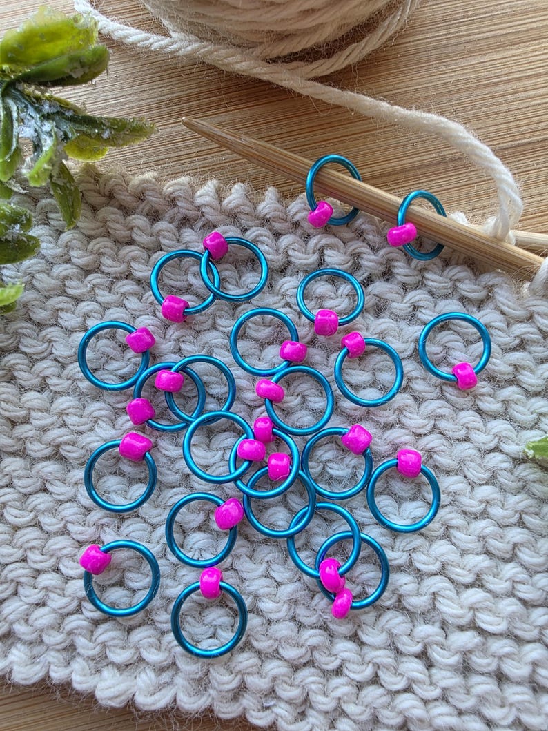 Pu&ograve; includere: Segnapunti color turchese con perline rosa sparsi su una superficie lavorata a maglia. Due segnapunti sono su un ferro da maglia di legno. Un gomitolo di lana e del verde sono sullo sfondo. Questi segnapunti sono per progetti di maglia e uncinetto.