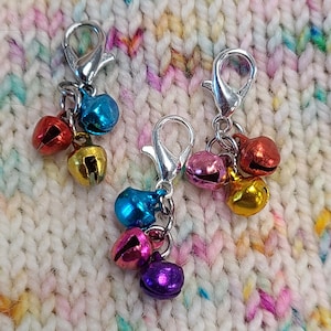 May include: Three silver stitch markers with colorful bells. Each marker has a silver clip and a chain with multiple bells in different colors, including blue, pink, red, gold, and purple.