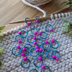 Pu&ograve; includere: Segnapunti turchese con perline rosa su un campione di maglia. Un gomitolo di lana color crema e ferri da maglia suggeriscono un progetto di lavoro a maglia. I segnapunti aiutano a tenere il conto delle maglie.