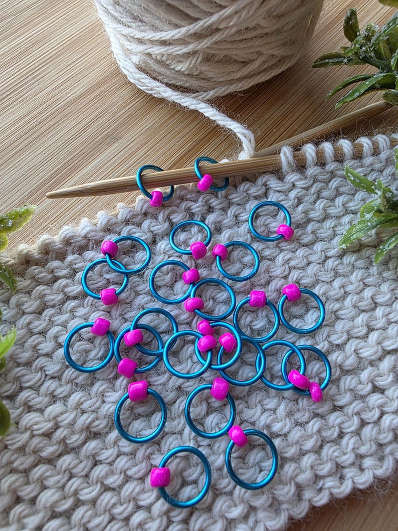 Pu&ograve; includere: Segnapunti turchese con perline rosa su un campione di maglia. Un gomitolo di lana color crema e ferri da maglia suggeriscono un progetto di lavoro a maglia. I segnapunti aiutano a tenere il conto delle maglie.