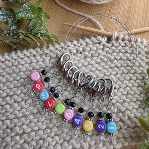 May include: A set of numbered stitch markers in various colors, attached to silver clasps. The markers are arranged on a beige knitted fabric, with a wooden knitting needle and a ball of yarn in the background. The numbers are from 1 to 9.