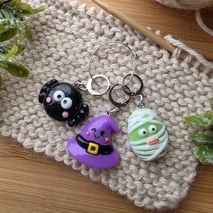 May include: Three Halloween-themed keychains: a black spider, a purple witch hat, and a green mummy. Each charm is attached to a silver keyring. The keychains are displayed on a beige knitted surface with a wooden knitting needle.