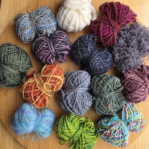 May include: A collection of various yarn balls in assorted colors and textures, including cream, dark green, blue, orange, purple, grey, and multi-colored strands. The yarn balls are arranged in a cluster, showcasing different fiber types and thicknesses.