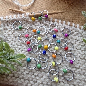 May include: A collection of silver-toned metal stitch markers, each featuring a single small, smooth, round bead in various vibrant colors including yellow, green, purple, orange, blue, pink, and red, scattered across a piece of beige knit fabric.