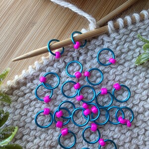 Pu&ograve; includere: Segnapunti turchesi con perline rosa a forma di cuore su tessuto a maglia. Un ferro da maglia in legno e un gomitolo di lana bianca suggeriscono un progetto di lavoro a maglia in corso.