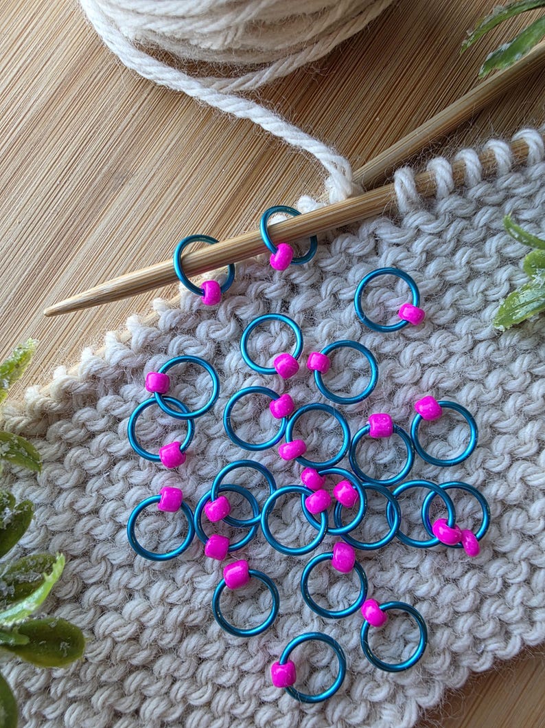 Pu&ograve; includere: Segnapunti turchesi con perline rosa a forma di cuore su tessuto a maglia. Un ferro da maglia in legno e un gomitolo di lana bianca suggeriscono un progetto di lavoro a maglia in corso.