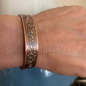 May include: A copper bracelet with an intricate swirl design. The bracelet is worn on a person's wrist.