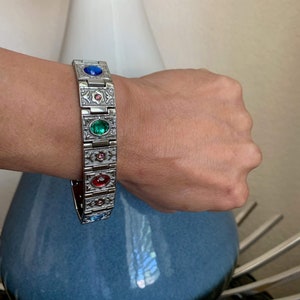 Peut inclure: Un bracelet en argent avec un motif décoratif et cinq pierres précieuses colorées. Les pierres précieuses sont rouge, bleu, vert, rose et bleu clair.