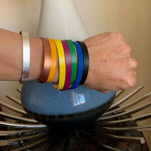 May include: A set of seven leather bracelets in various colors, including silver, copper, yellow, green, purple, blue, and black. The bracelets are stacked on a person's wrist.