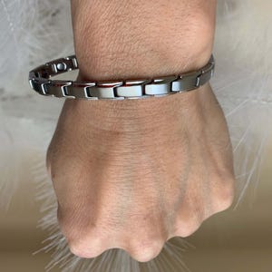 May include: A silver magnetic bracelet with a clasp closure. The bracelet is made of multiple linked metal pieces.