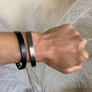 May include: Two bracelets on a wrist. One is a black leather bracelet with a silver buckle. The other is a silver metal cuff bracelet.