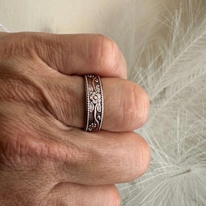 May include: A silver-colored ring with an engraved floral pattern. The ring features a repeating design of flowers and leaves, with a beaded border. The ring is worn on a finger.