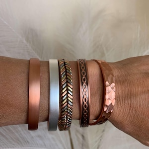May include: A collection of five metal bracelets in various designs and finishes. The bracelets include a smooth copper band, a silver band, a braided band with copper, silver, and gold tones, a patterned copper band, and a hammered copper band.