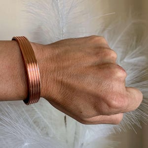 Op de afbeelding: Een koperkleurige armband met een gestapeld ontwerp. De armband is gemaakt van meerdere, dunne metalen banden, waardoor een textuur ontstaat. De armband wordt om een pols gedragen en toont zijn eenvoudige maar elegante ontwerp.