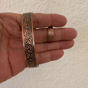 Op de afbeelding: Twee koperen armbanden met een ingewikkeld swirl-ontwerp. De grotere armband is breder dan de kleinere.