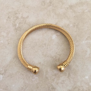 Puede incluir: Un brazalete dorado con un diseño trenzado y extremos redondeados.