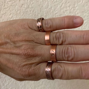 May include: Four copper rings of different designs are shown on fingers. One ring is plain, another has a heart, and two feature detailed patterns. The rings are a warm, reddish-brown colour.