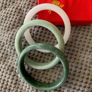 May include: Three jade bangles, one white and two green, stacked on top of each other. The bangles are on a gray textured surface.