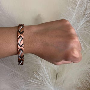 May include: A copper bracelet with a braided design and clear crystals. The bracelet is on a person's wrist.