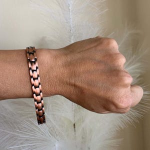 Peut inclure: Bracelet de couleur cuivre avec un design à maillons rectangulaires. Le bracelet est porté au poignet, mettant en valeur son éclat métallique. Les maillons sont un mélange de cuivre et de tons plus foncés, créant un aspect texturé. Le fond est de couleur neutre.