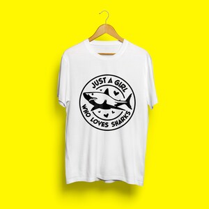 May include: White t-shirt with a black circle graphic featuring a shark and the text "Just a girl who loves sharks".