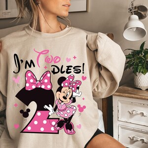 May include: Beige sweatshirt featuring a Minnie Mouse graphic with the text "I'm Two Dles!". The design includes a large number 2, a bow, hearts, and Minnie Mouse in a pink polka dot dress. A fun and festive design for a child's birthday.
