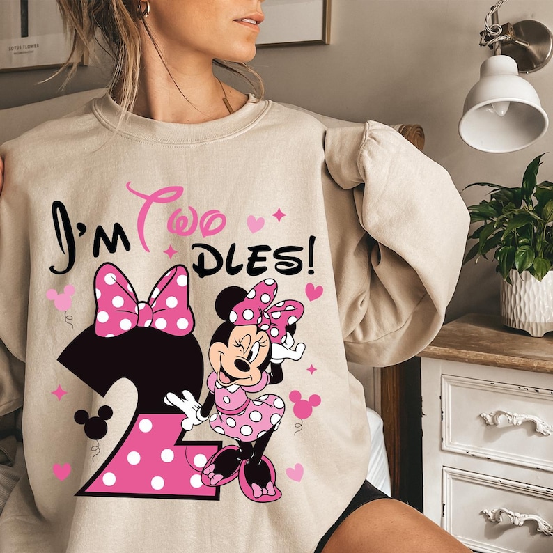 May include: Beige sweatshirt featuring a Minnie Mouse graphic with the text "I'm Two Dles!". The design includes a large number 2, a bow, hearts, and Minnie Mouse in a pink polka dot dress. A fun and festive design for a child's birthday.