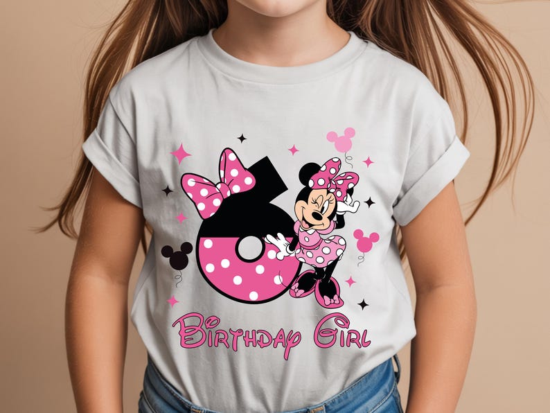 May include: A light gray t-shirt featuring a Minnie Mouse graphic. The design includes a large number 6 with pink polka dots, a bow, and the words "Birthday Girl" in pink script. Minnie Mouse is also featured.