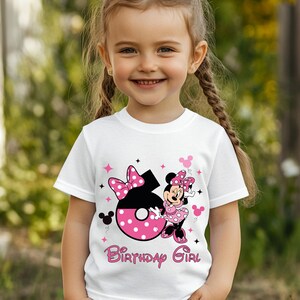 May include: White t-shirt featuring a Minnie Mouse design with a large number 6 in black and pink polka dots. The shirt has the words "Birthday Girl" in pink script. Pink stars and Minnie Mouse silhouettes decorate the shirt.