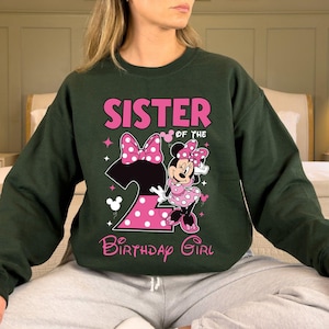 Puede incluir: Sudadera verde oscuro con el texto "SISTER OF THE BIRTHAY GIRL" en rosa. El dise&ntilde;o incluye un gran n&uacute;mero 2 rosa con lunares blancos, un lazo rosa y un gr&aacute;fico de Minnie Mouse con un vestido rosa de lunares.