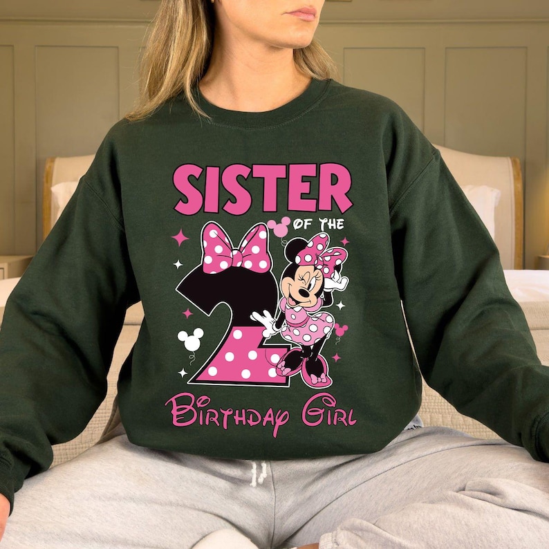 Puede incluir: Sudadera verde oscuro con el texto "SISTER OF THE BIRTHAY GIRL" en rosa. El dise&ntilde;o incluye un gran n&uacute;mero 2 rosa con lunares blancos, un lazo rosa y un gr&aacute;fico de Minnie Mouse con un vestido rosa de lunares.