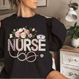 May include: Black sweatshirt with a floral design and the word "NURSE" in a decorative font. The design includes a nurse's cap with a red cross, a stethoscope, and a pink heart. The sweatshirt is a comfortable, casual style.