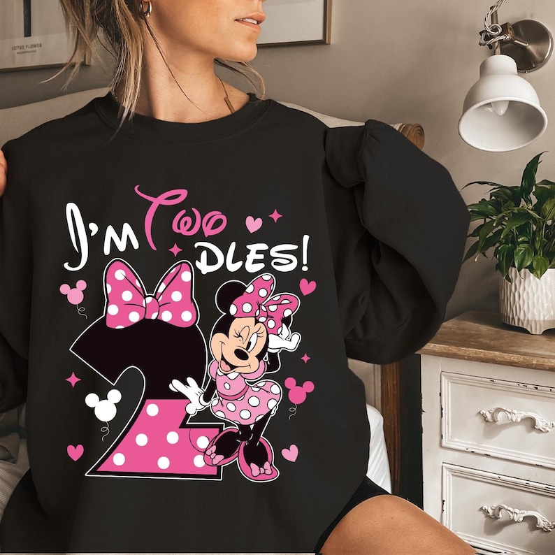 May include: Black sweatshirt featuring Minnie Mouse with the text "I'm Two Dles!" in white and pink. The design includes a large pink polka dot bow, the number 2, and Minnie Mouse in a pink polka dot dress.