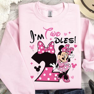 Może przedstawiać: Różowa bluza z napisem "I'm Two Dles!" i grafiką Myszki Minnie. Cyfra "2" jest różowa w białe kropki. Projekt zawiera serca i gwiazdki.