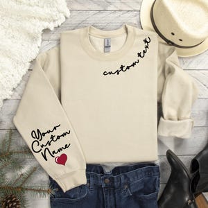 May include: A cream-colored sweatshirt with the words "custom text" and "Your Custom Name" in black script. A red heart graphic is below the name on the sleeve. The sweatshirt is folded on a wooden surface with a hat and boots.