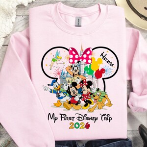 May include: A light pink sweatshirt featuring a Disney-themed graphic. The design includes Mickey and Minnie Mouse, Donald Duck, Goofy, and Pluto. The text "My First Disney Trip 2026" is displayed below the characters, with the name "Helena" above.